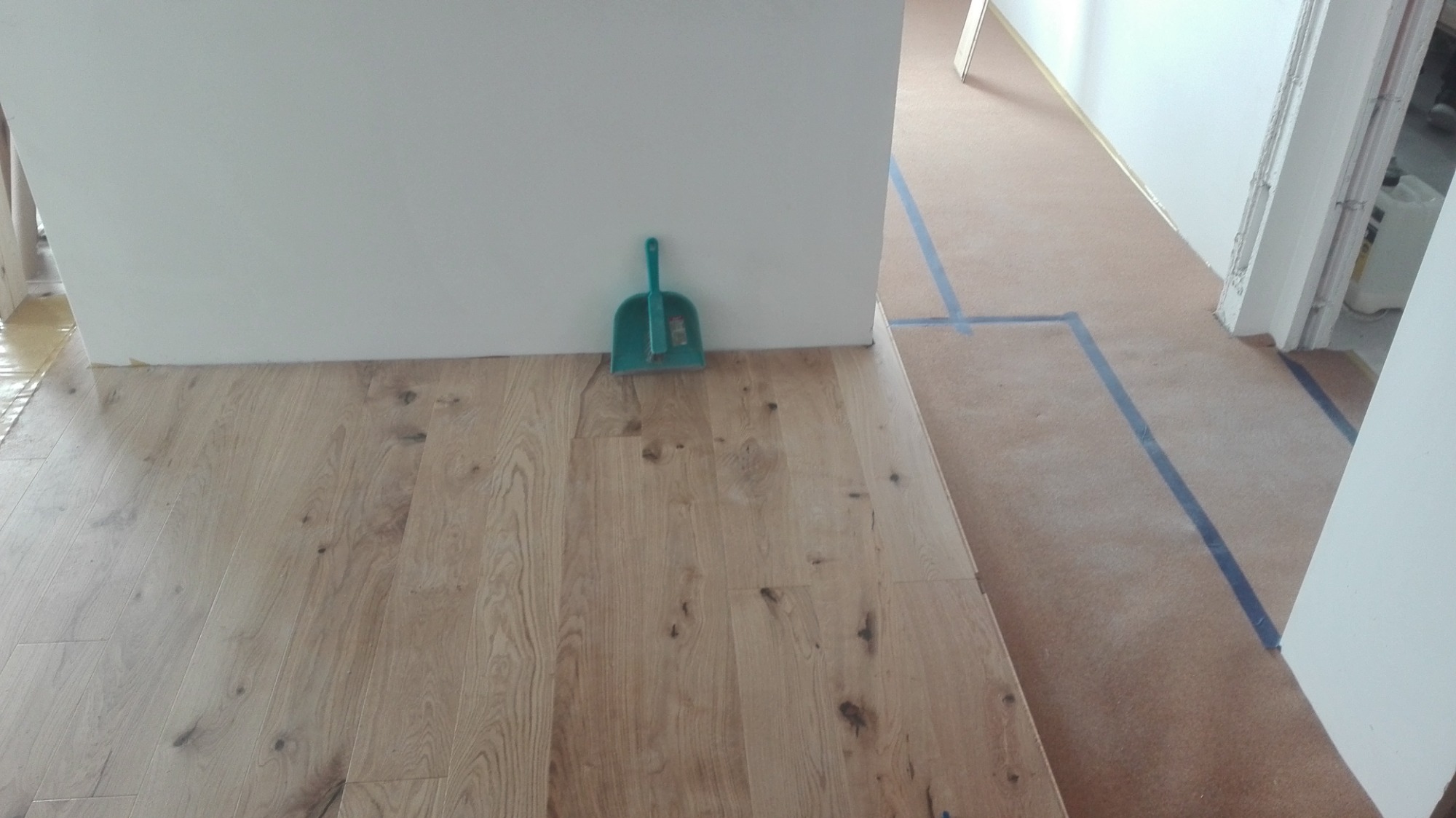 Układanie jasnej drewnianej podłogi, widoczna niebieska taśma malarska na zabezpieczonej podłodze w tle i zielona zmiotka przy ścianie.