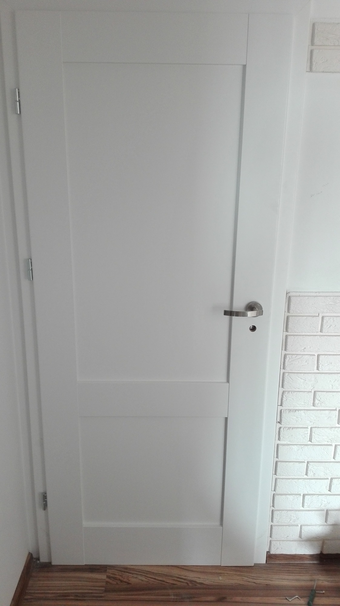 Białe, dwupanelowe drzwi wewnętrzne z srebrną klamką i widoczną ścianą wykończoną cegłą dekoracyjną.