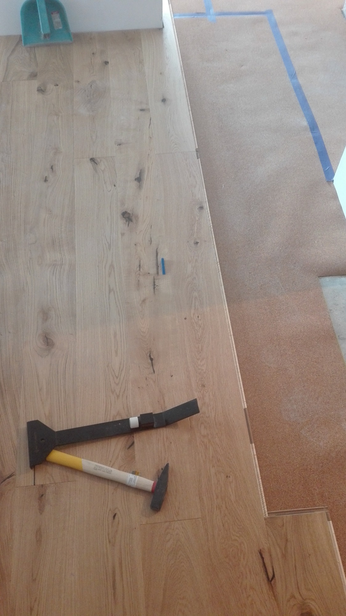 Montaż drewnianej podłogi: widoczne panele, podkład korkowy, niebieska taśma malarska, dobijak i młotek.
