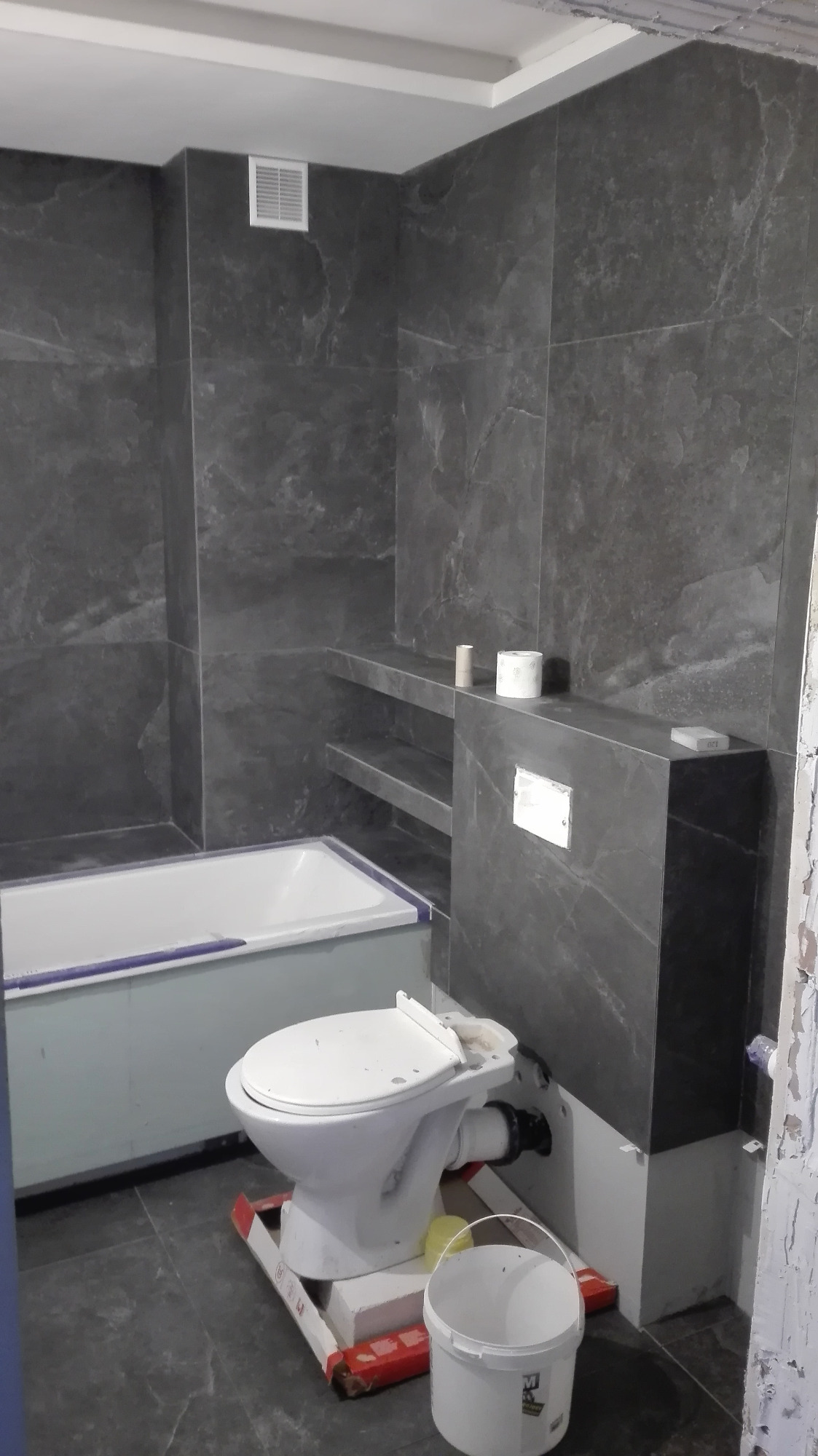 Łazienka w trakcie remontu: szare płytki imitujące marmur na ścianach, częściowo zamontowana toaleta na podwyższeniu, wanna z obudową, widoczne narzędzia i wiadro, surowy stan wykończenia.