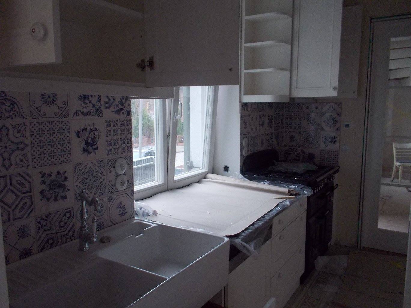 Kuchnia w trakcie prac wykończeniowych: białe szafki wiszące, ozdobne płytki ceramiczne w odcieniach błękitu i bieli na ścianie nad zlewem, zabezpieczony blat roboczy folią, widoczny fragment...