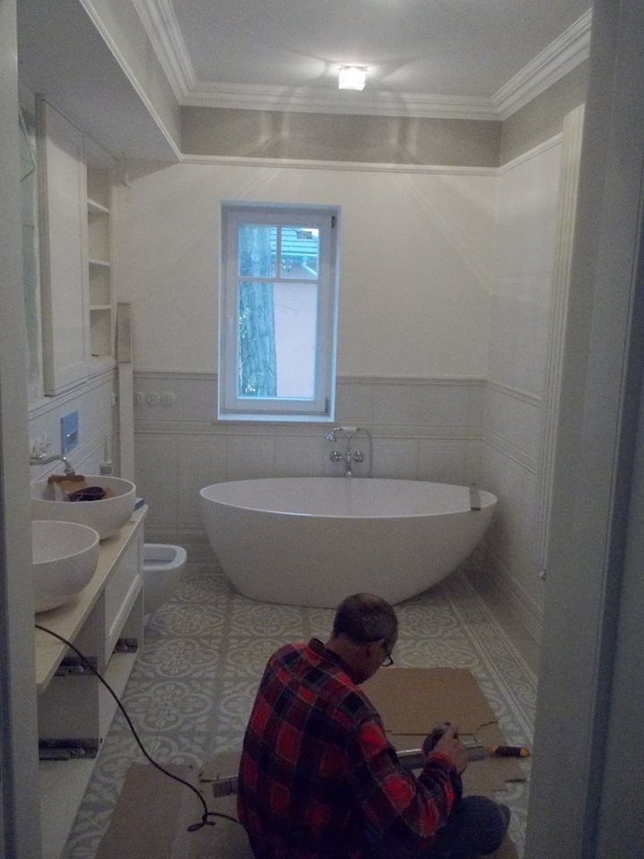 Instalacja paneli podłogowych w łazience z wolnostojącą wanną i umywalkami nablatowymi; pracownik w koszuli w kratę przycina panel na podłodze o wzorzystym deseniu.