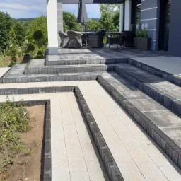 Nowoczesny taras i ścieżka z kostki brukowej w odcieniach szarości i beżu, prowadząca do domu z kolumnami i meblami ogrodowymi.