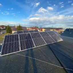 Rząd paneli słonecznych na płaskim dachu z bitumicznej papy, widok z góry na osiedle domów jednorodzinnych pod błękitnym niebem z chmurami.