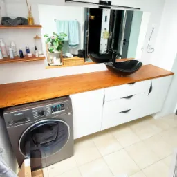 Łazienka z drewnianym blatem nad pralką, białe szafki z trzema szufladami i czarna umywalka nablatowa w kształcie misy. Na ścianie lustro z oświetleniem. Na półkach kosmetyki i dekoracje.