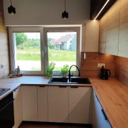 Nowoczesna kuchnia z białymi szafkami, drewnianym blatem i czarną armaturą, widok z okna na zielony ogród, oświetlenie LED podszafkowe.