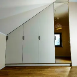 Biała szafa wnękowa z czterema drzwiami, dopasowana do skosu poddasza, z czarnymi, podłużnymi uchwytami, na drewnianej podłodze, widok na pokój z oknem zza szafy.