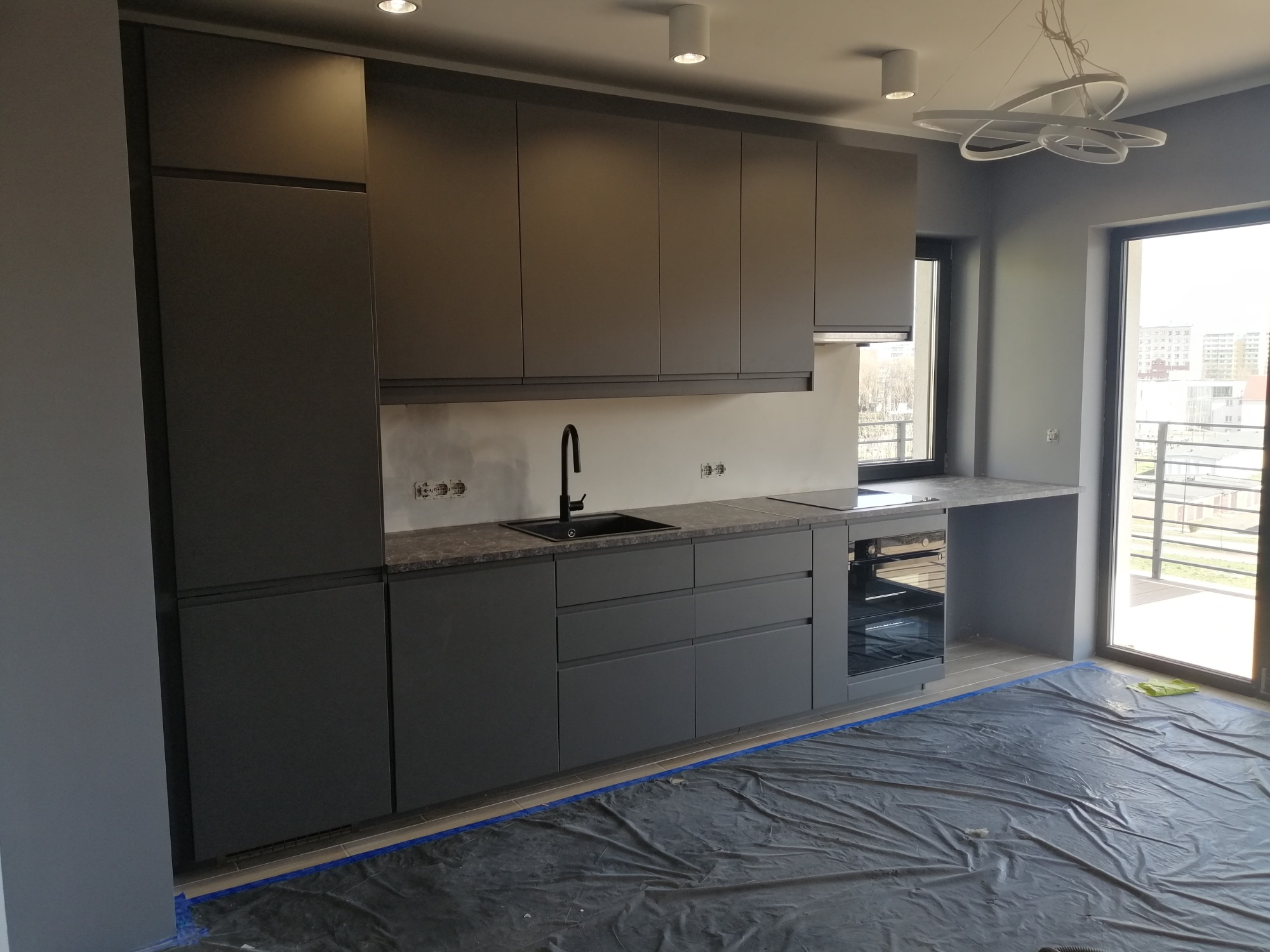 Szara kuchnia na wymiar z czarną armaturą, blatem imitującym kamień i płytą indukcyjną, widok na balkon zza okna, podłoga zabezpieczona folią malarską.
