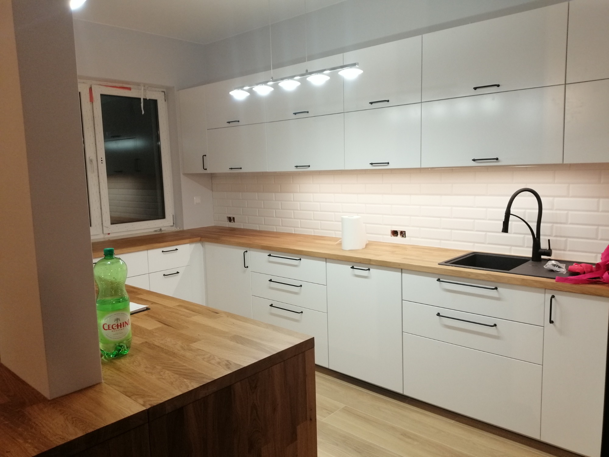 Nowoczesna, jasna kuchnia z białymi szafkami z minimalistycznymi, czarnymi uchwytami i drewnianym blatem, oświetlona listwą lamp nad szafkami, widoczna butelka wody mineralnej na wyspie kuchennej.