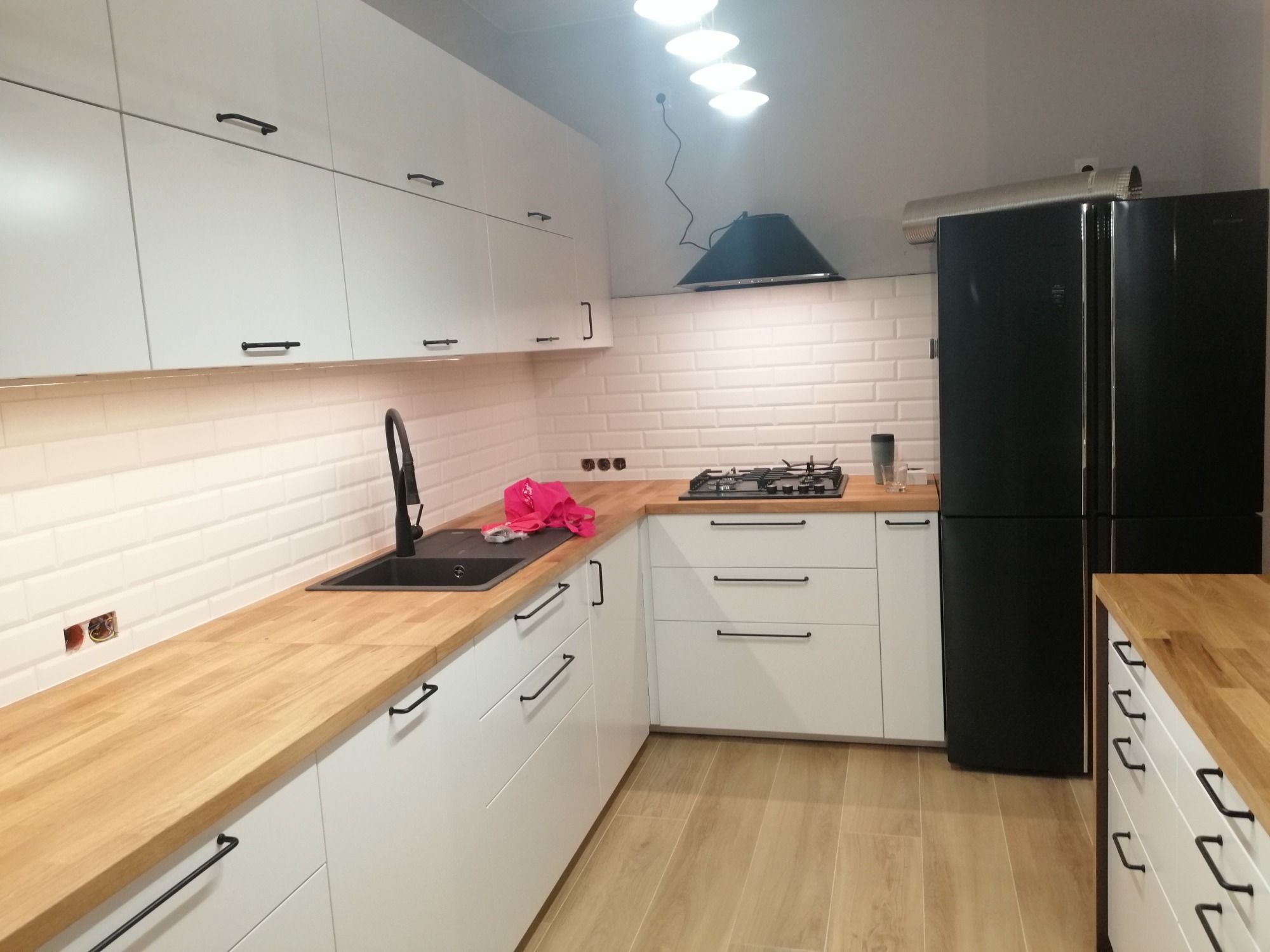 Nowoczesna kuchnia z białymi szafkami i drewnianym blatem, czarną armaturą i lodówką w stylu retro, widoczne gniazdka elektryczne w ścianie z białej cegły.