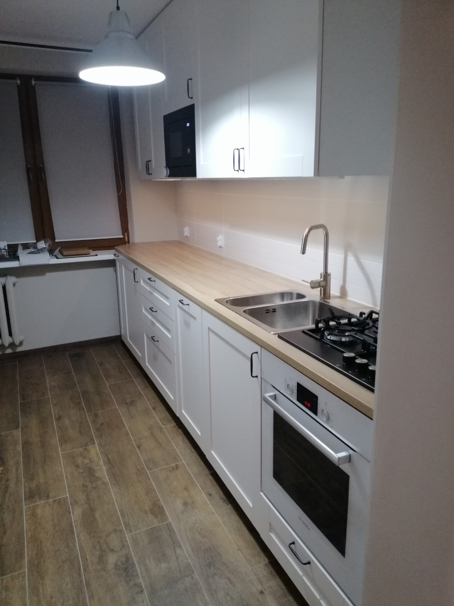 Biała kuchnia z drewnianym blatem, widok na zlew, płytę gazową i piekarnik. Nad blatem wiszą białe szafki z czarnymi uchwytami.