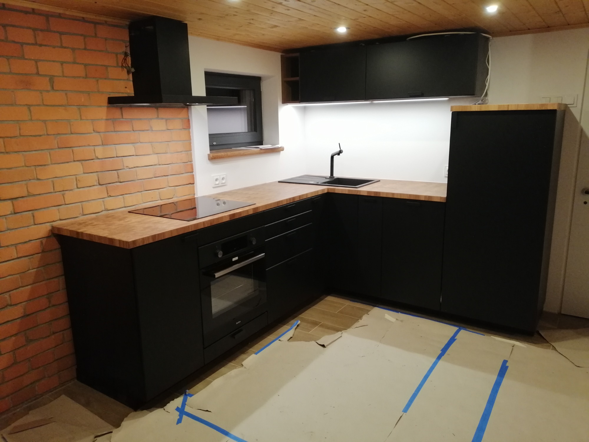 Częściowo ukończona kuchnia: czarne szafki z drewnianymi blatami, ceglana ściana, zabezpieczona podłoga kartonem i taśmą malarską.