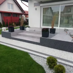Schody wejściowe do domu wyłożone szarymi płytkami, otoczone kostką brukową w jasnych odcieniach, z donicami z kwiatami i ozdobnymi trawami, widok na zadbany trawnik.