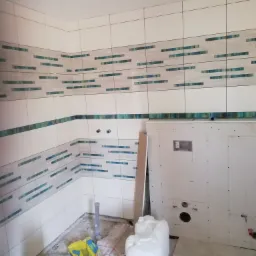 Białe płytki z poziomymi, turkusowymi paskami układane na ścianach w rogu pomieszczenia, widoczne przygotowania do instalacji sanitarnej i płyta kartonowo-gipsowa.