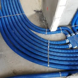 Niebieskie rury wentylacyjne karbowane, spięte metalowymi taśmami, prowadzone w surowym stanie podłogi budynku w trakcie instalacji rekuperacji.