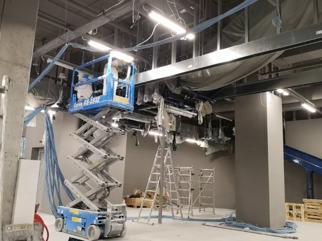 Prace instalacyjne w Dynowie: podnośnik nożycowy Genie GS-1932 w trakcie montażu instalacji pod sufitem, widoczne kable i rusztowania.