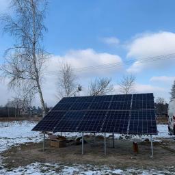 MILTON-PV - Panele słoneczne na metalowej konstrukcji, częściowo pokryte śniegiem, z kartonowym pudłem pod spodem, na tle błękitnego nieba z chmurami i brzozy bez liści.