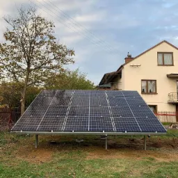 Wolnostojąca instalacja fotowoltaiczna na metalowej konstrukcji, umieszczona na trawniku przed domem jednorodzinnym w pochmurny dzień.