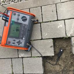 MILTON-PV - Miernik rezystancji uziemienia MRU-21 marki Sonel z wyświetlanym wynikiem 987 omów, umieszczony na szarej kostce brukowej, z czarnym przewodem wpiętym w ziemię.