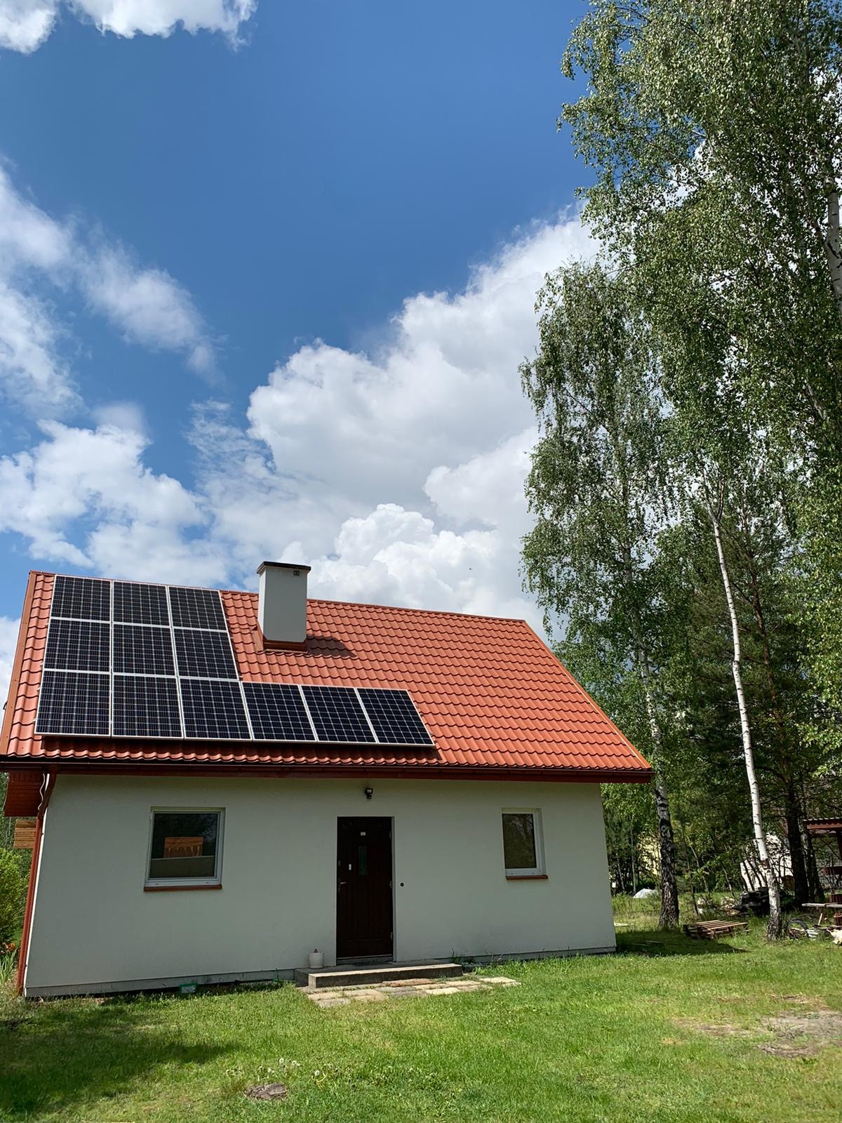 Dom jednorodzinny z panelami słonecznymi na czerwonym dachu, błękitne niebo z białymi obłokami w tle, drzewa po prawej stronie.