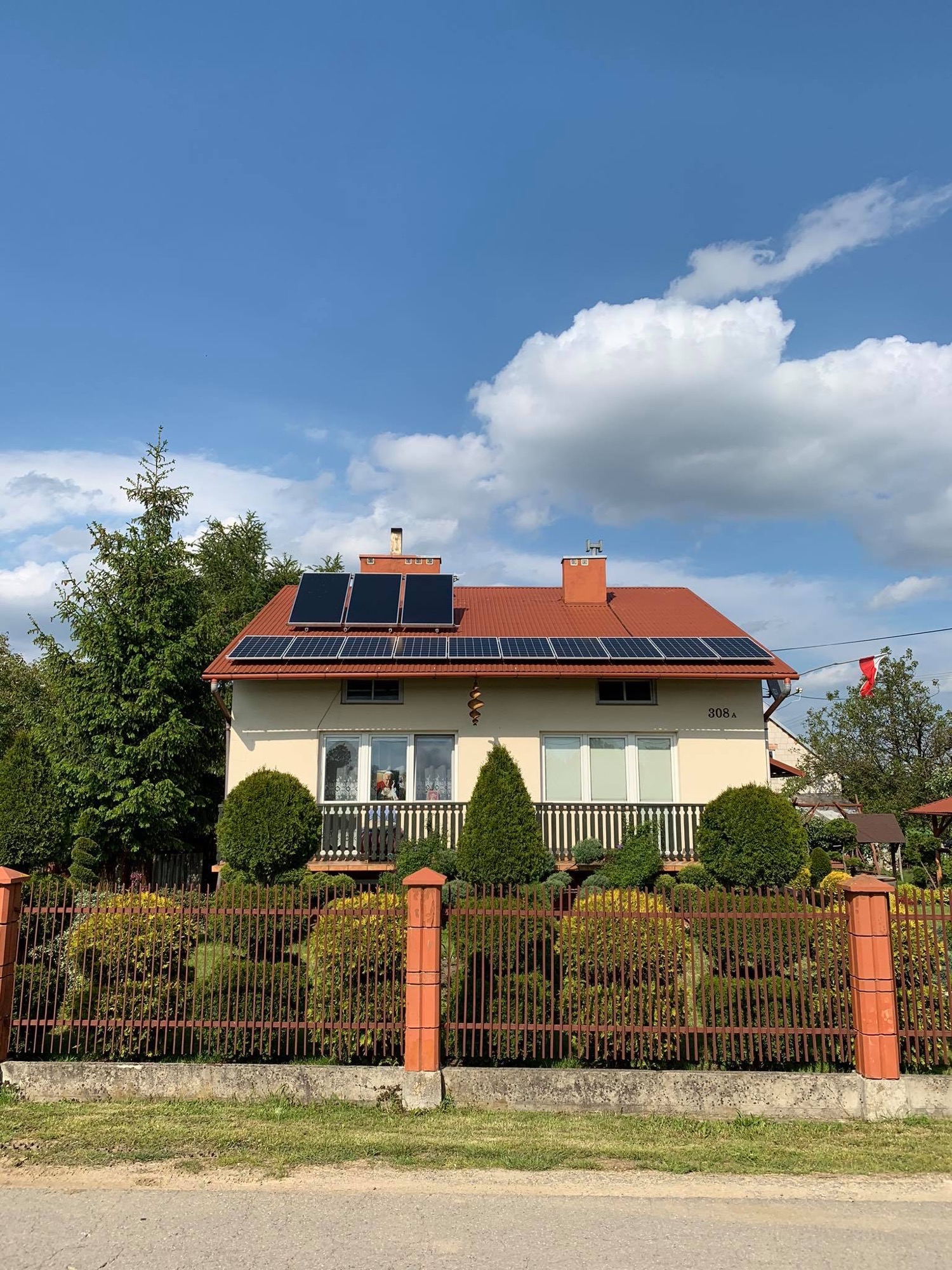 Dom jednorodzinny z czerwonym dachem i zainstalowanymi panelami słonecznymi na dachu, widok zza ogrodzenia.