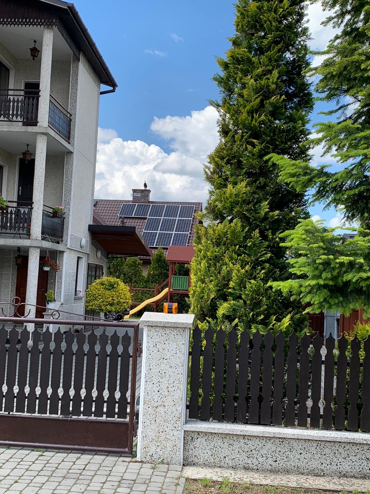 Dom jednorodzinny z brązowym dachem, na którym zamontowano panele słoneczne; w tle widoczny plac zabaw dla dzieci.