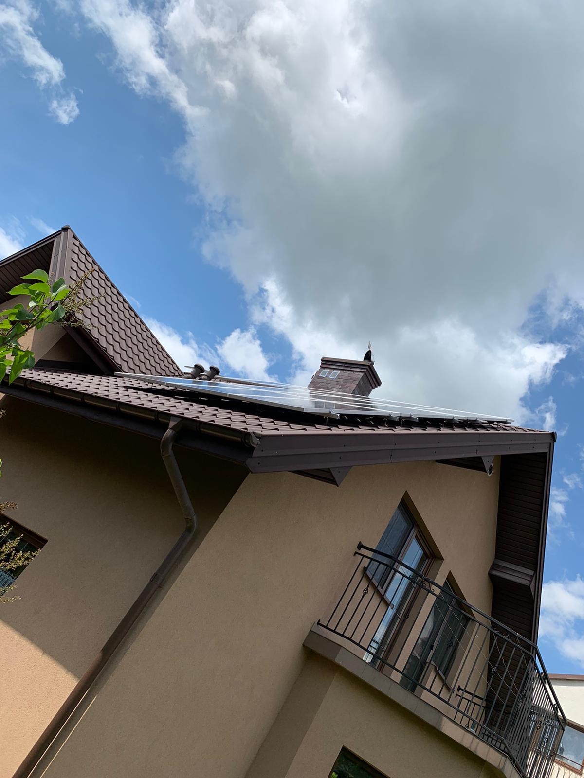 Instalacja paneli fotowoltaicznych na brązowym dachu domu jednorodzinnego z widocznym kominem i błękitnym niebem z chmurami.