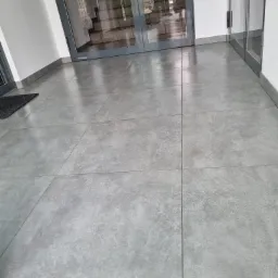 Szare płytki podłogowe wewnątrz budynku, widok na przeszklone drzwi wejściowe i schody w tle.