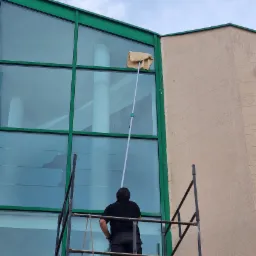 Pracownik na rusztowaniu myje okna budynku biurowego za pomocą mopa na długim kiju. Widoczne duże okna z zielonymi ramami i fragment elewacji budynku.