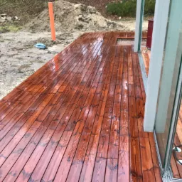 Świeżo zaimpregnowany, mokry drewniany podest tarasu z widoczną konstrukcją przesuwnych drzwi balkonowych i przygotowanym podłożem pod trawnik.