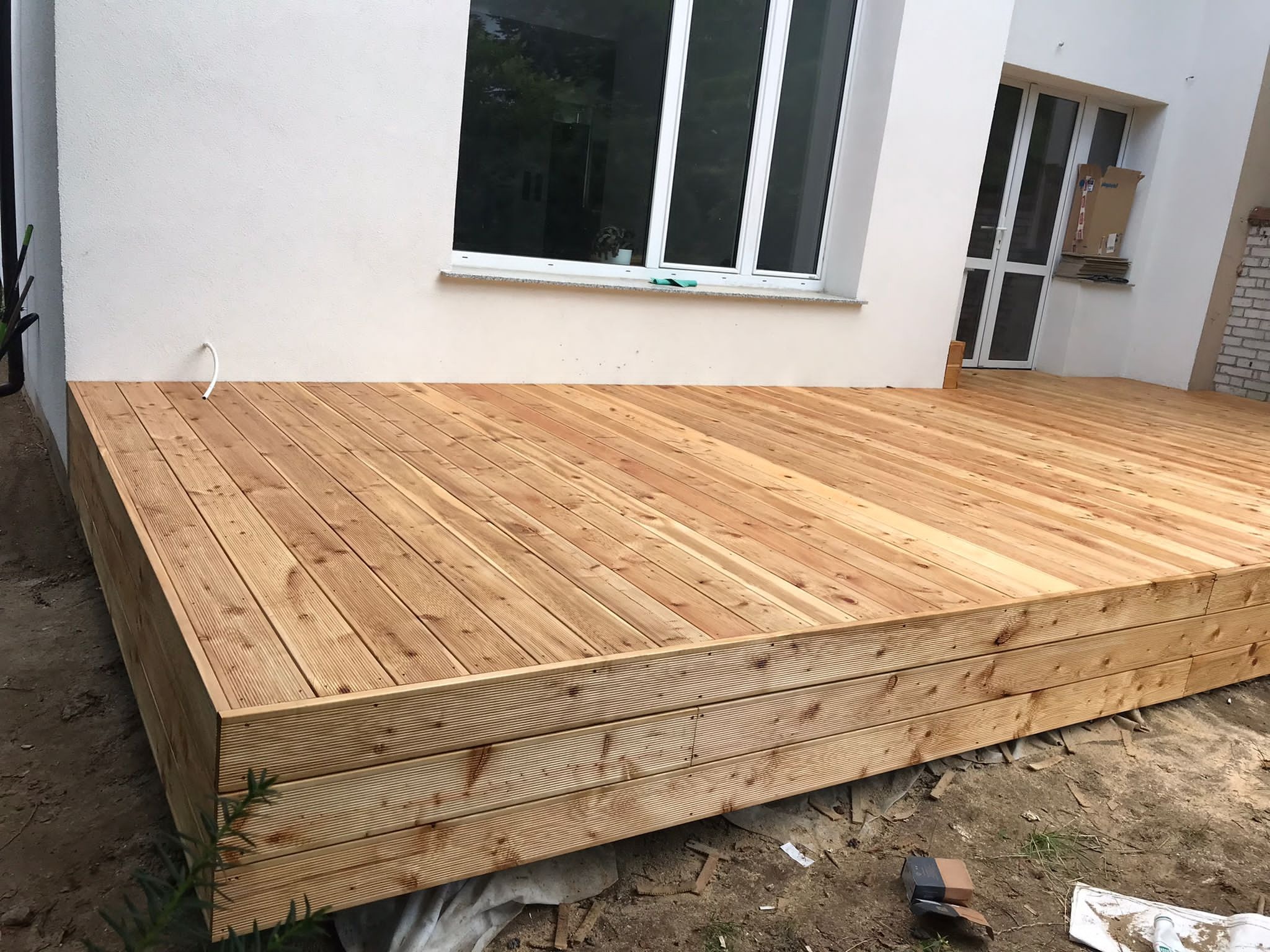 Nowoczesny drewniany podest przydomowy z desek ryflowanych, widok z boku, z białymi ścianami budynku w tle i oknami.