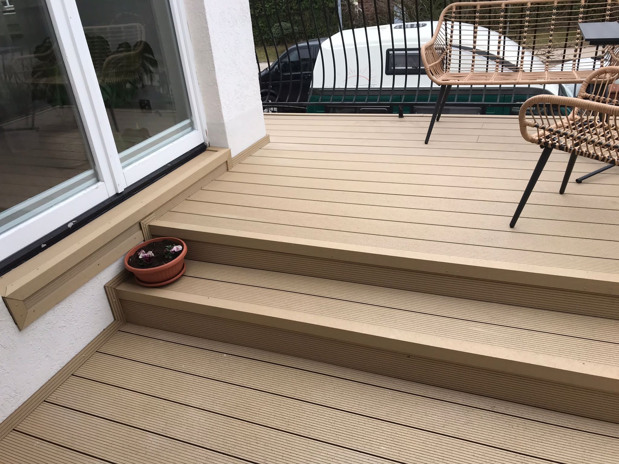 Jasny taras z kompozytu z dwoma stopniami, wiklinowe krzesła w tle i doniczka z kwiatami przy drzwiach tarasowych.