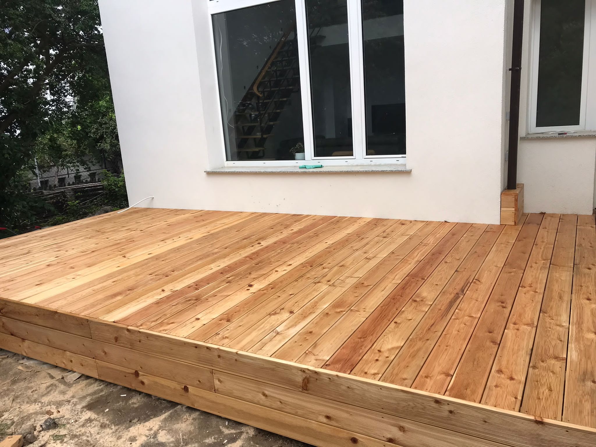 Nowa drewniana konstrukcja tarasu przylegająca do białego budynku z widocznymi schodami wewnątrz przez okno.