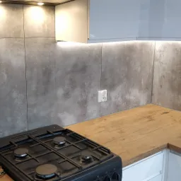 Nowoczesna kuchnia z szarymi płytkami imitującymi beton, drewnianym blatem, białą zabudową i czarną płytą gazową. Na blacie stoi butelka z pojedynczym tulipanem.