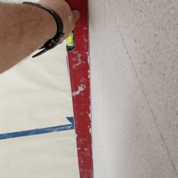 Tomasz Mazur - Czerwona poziomica przyłożona do szorstkiej ściany podczas prac wykończeniowych, widoczna ręka z czarnym zegarkiem, niebieska taśma malarska na podłodze.