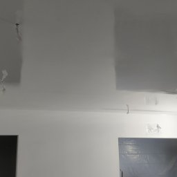 Tomasz Mazur - Świeżo pomalowany sufit na wysoki połysk, z widocznymi foliami ochronnymi na elementach oświetlenia, oraz zabezpieczonymi folią otworami drzwiowymi, w jasnym pomieszczeniu.