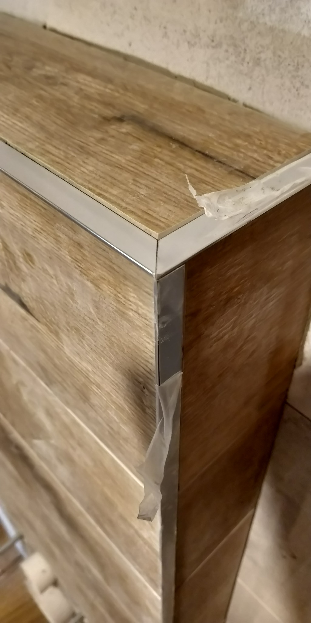 Fragment szafki wykończonej płytkami imitującymi drewno, zabezpieczonej srebrną listwą wykończeniową, z resztkami folii ochronnej na krawędziach.