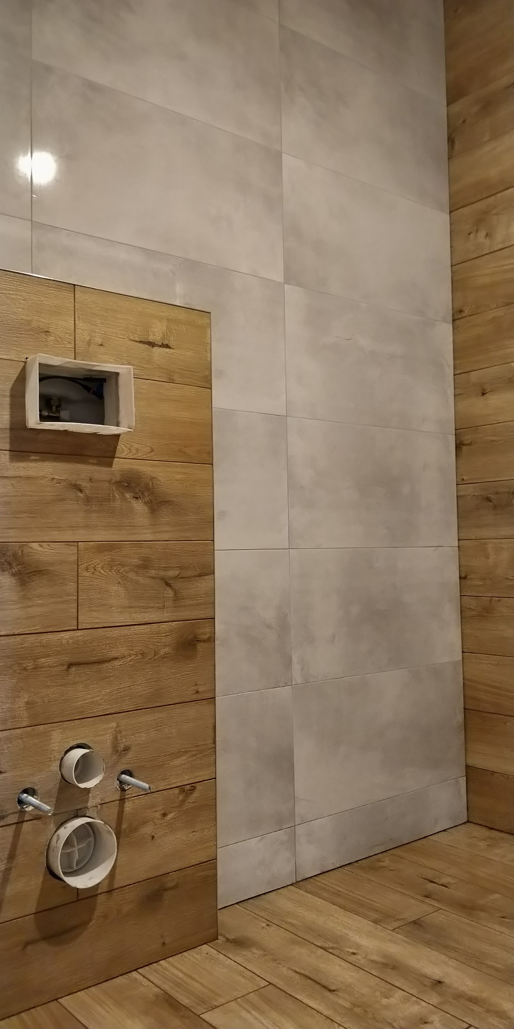 Wnętrze łazienki w trakcie remontu: połączenie płytek imitujących drewno i szarych, matowych płytek ściennych, widoczne rury i instalacja podtynkowana.