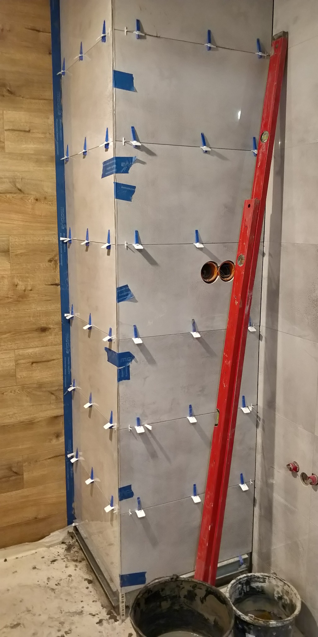 Świeżo ułożone szare płytki na ścianie z widocznymi krzyżykami dystansowymi, poziomica oparta o ścianę, przygotowane wiaderka z zaprawą i wystające z ściany rury instalacji wodnej.