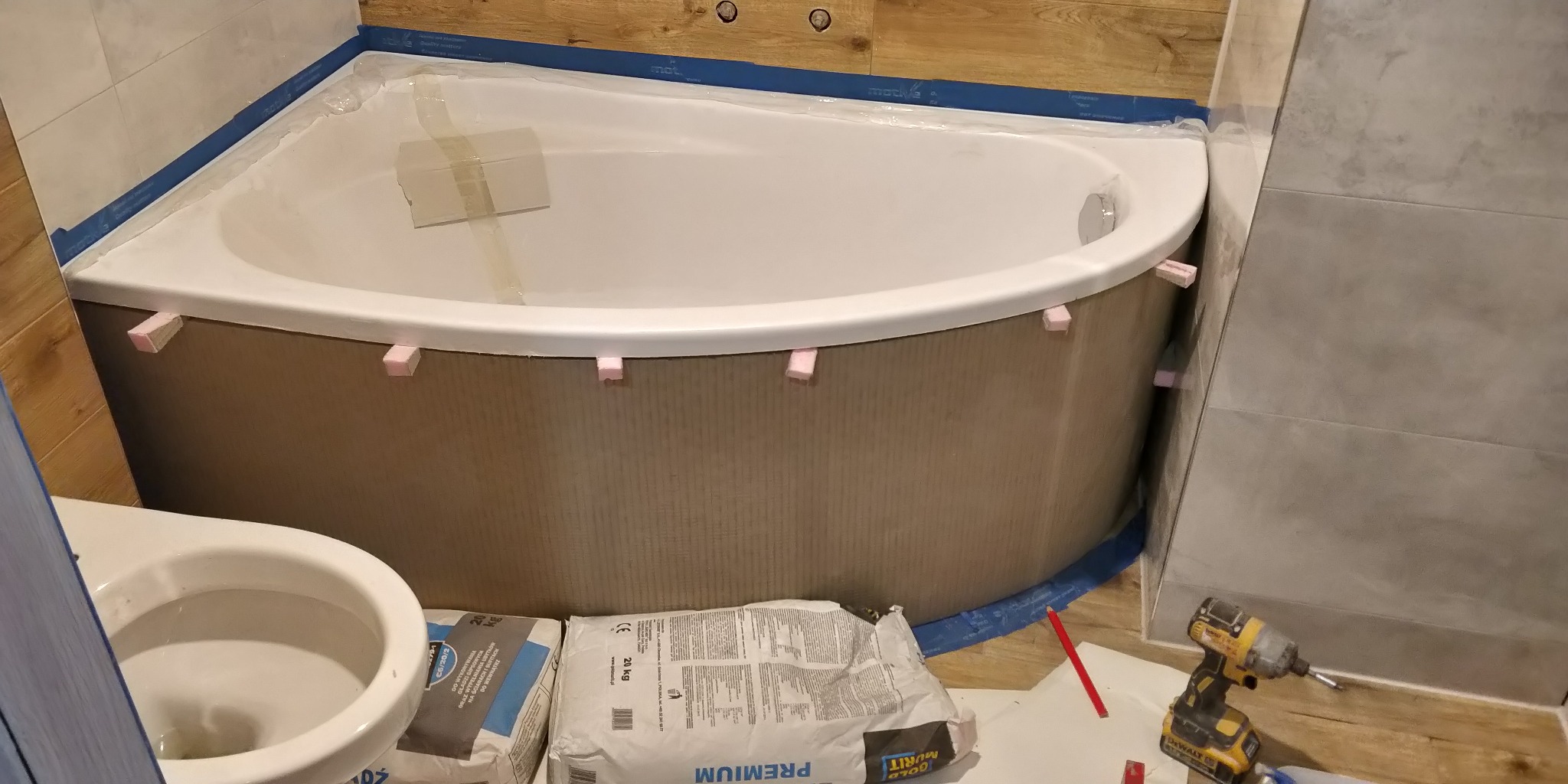 Montaż obudowy wanny narożnej z użyciem piankowych rozpórek i taśmy maskującej, widoczne worki z zaprawą klejową i narzędzia, w tle wykończone płytki ścienne.