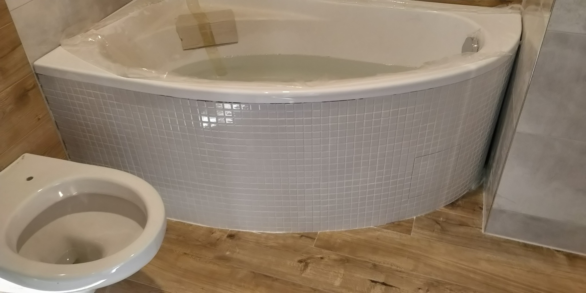 Nowa, narożna wanna obudowana mozaiką z małych, białych płytek, częściowo zabezpieczona folią ochronną, obok biała muszla klozetowa, drewnopodobna podłoga i szare płytki ścienne.