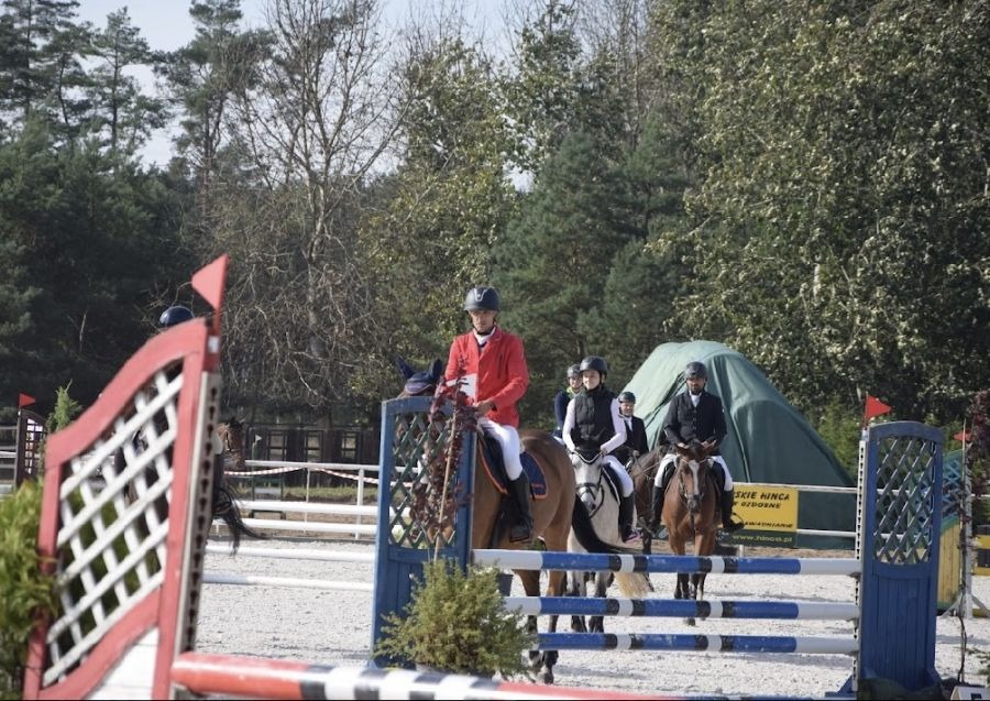 Grupa jeźdźców na koniach w strojach sportowych, oczekujących na przejazd przez przeszkodę z czerwono-białymi elementami, na tle drzew i namiotu.