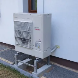 Biała zewnętrzna jednostka klimatyzacji Atlantic na metalowej konstrukcji, zamontowana przy białej ścianie budynku z brązowym oknem, widoczna trawa i kostka brukowa.