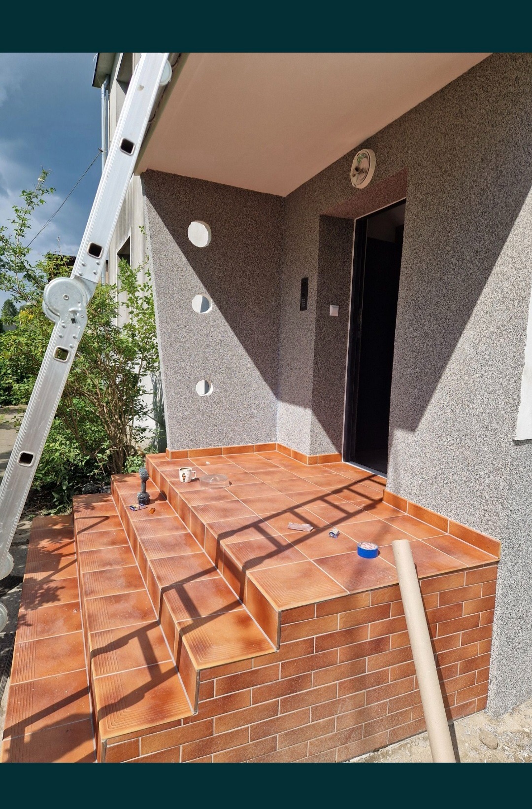 Wyłożone terakotą schody wejściowe do domu z elewacją w kolorze szarego baranka, z widoczną drabiną opartą o budynek, narzędziami i kubkiem na schodach.