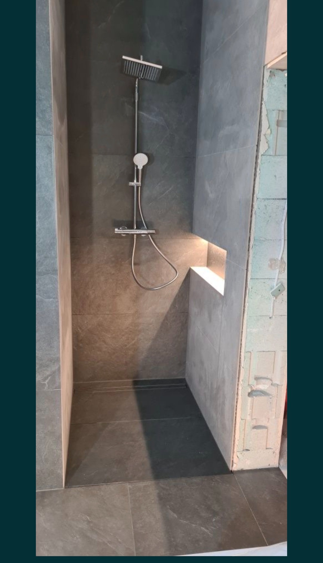 Wykończona kabina prysznicowa z szarymi płytkami imitującymi kamień, wyposażona w deszczownicę i oświetloną wnękę na kosmetyki.