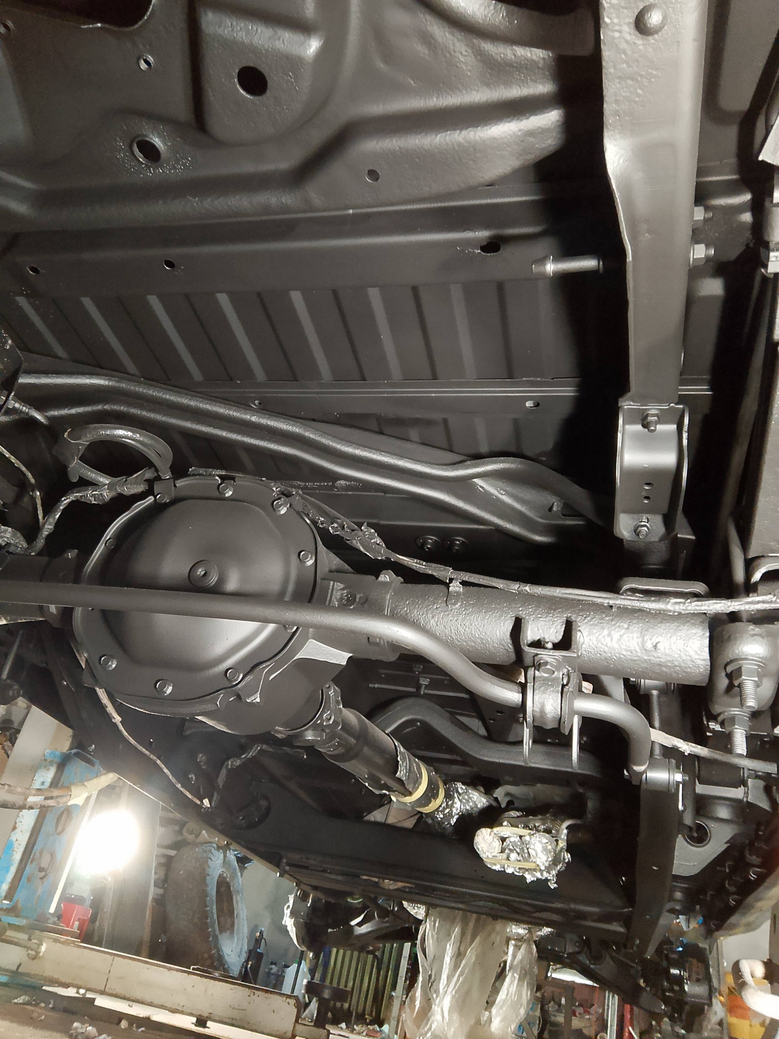 Podwozie samochodu po zabezpieczeniu antykorozyjnym, widok od spodu. Elementy zawieszenia i układu napędowego pokryte czarną farbą ochronną. Widoczne detale konstrukcyjne.