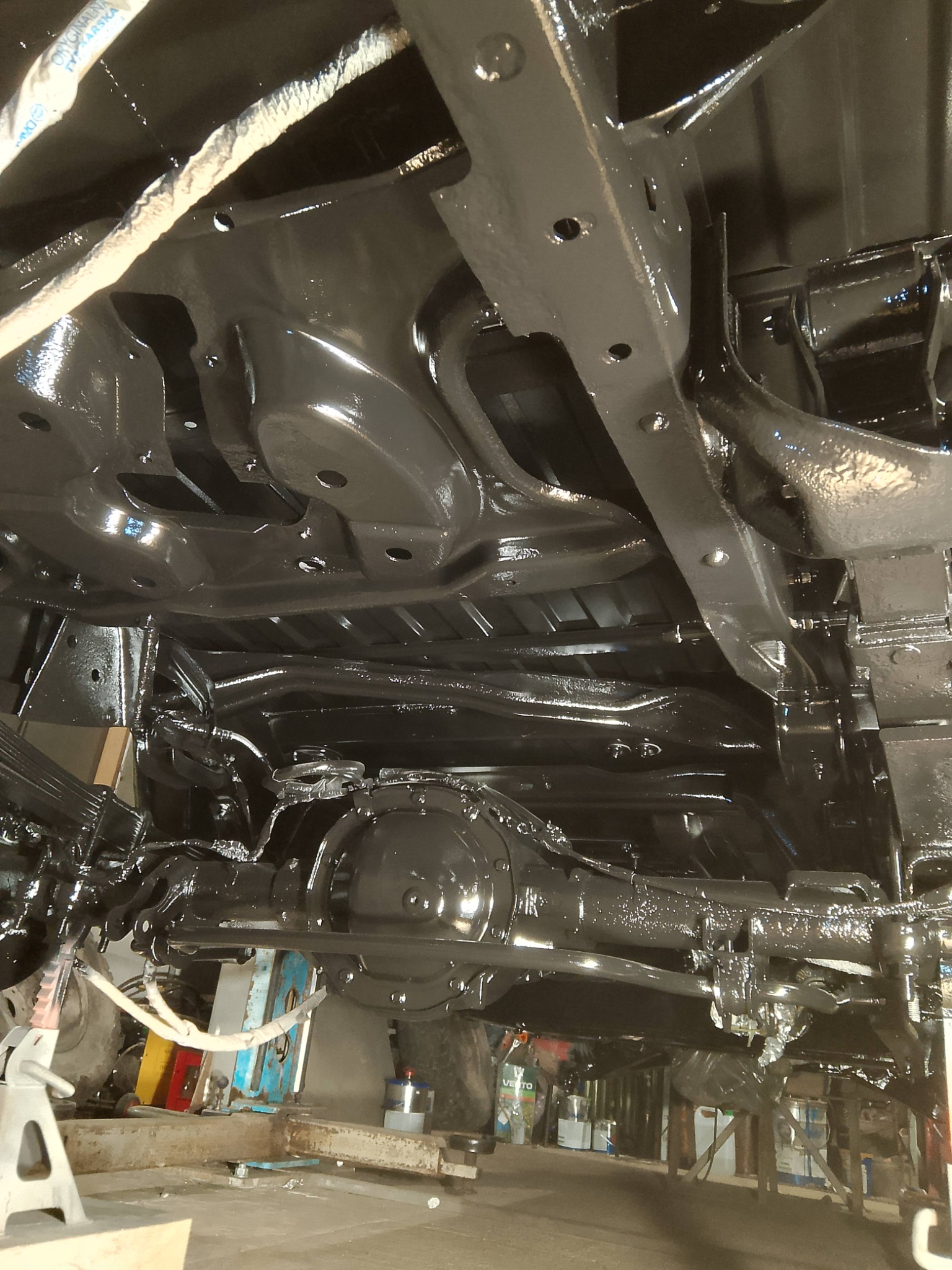 Podwozie samochodu po zabezpieczeniu antykorozyjnym, widok od spodu. Elementy zawieszenia i ramy pokryte czarną, błyszczącą powłoką.