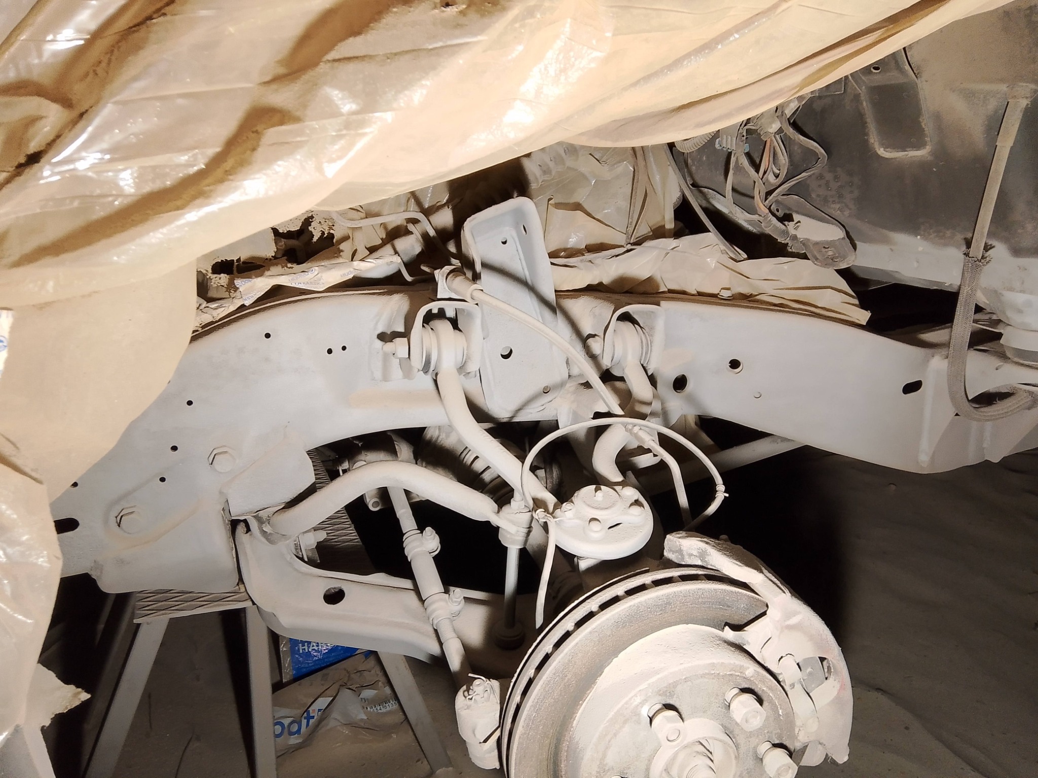 Podwozie samochodu po konserwacji antykorozyjnej, zabezpieczone folią. Widoczne elementy zawieszenia i tarczy hamulcowej w jasnym kolorze. Detale starannie pokryte.