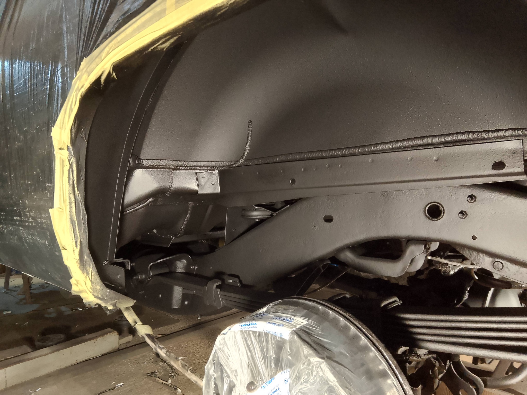 Częściowo owinięte folią i taśmą podwozie samochodu po zabezpieczeniu antykorozyjnym, pomalowane na czarno. Widoczne elementy zawieszenia i ramy.