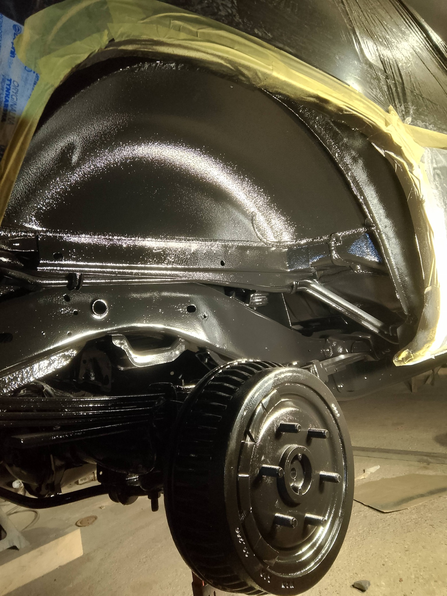 Podwozie samochodu po zabezpieczeniu antykorozyjnym, widoczne elementy zawieszenia i błyszcząca, czarna powłoka ochronna. Zabezpieczone taśmą elementy.